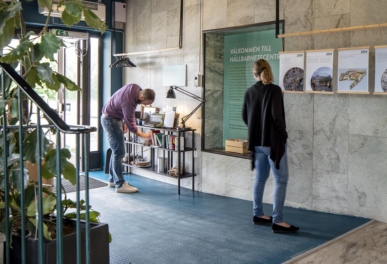 Två personer i entrén dekorerad med infoposter, böcker och andra småsaker. Foto