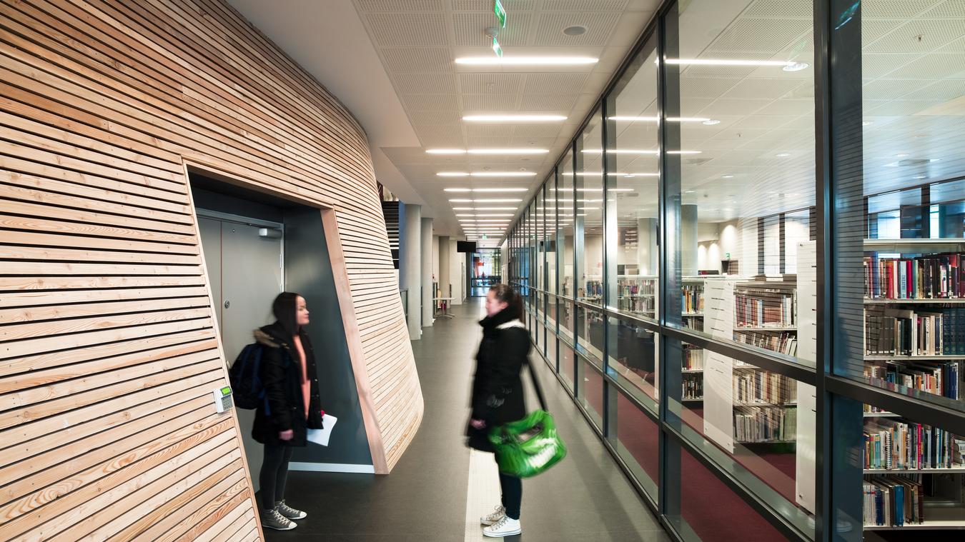 Hall med trävägg och glasfasad samt två elever. Foto