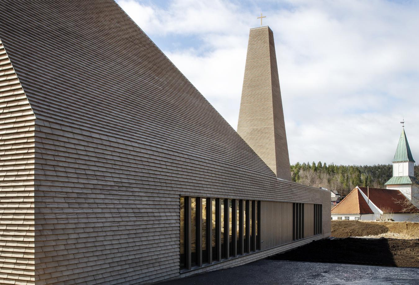 Ny kyrkobyggnad med gammal kyrka i bakgrunden. Foto